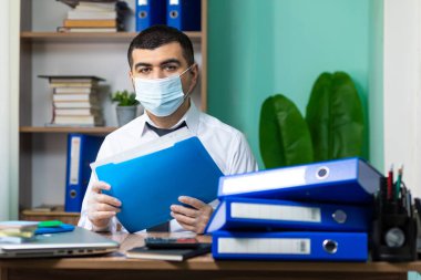 Genç işadamı masada oturuyor, elinde belgelerle tıbbi koruyucu maske ve eldivenlerle kameraya bakıyor.