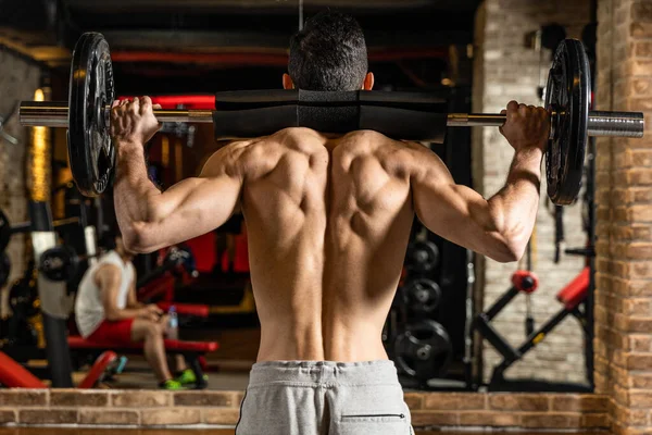 Back view of young bodybuilder flexing muscles with barbell in front of mirror at gym