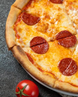 Close up of pepperoni pizza with melted mozzarella cheese being served on a dark background with a fresh tomato