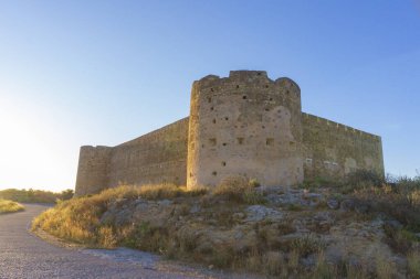 Gün batımında tepede güçlü kuleleri ve garaj yolu olan etkileyici bir ortaçağ kalesi, açık mavi gökyüzüne karşı..