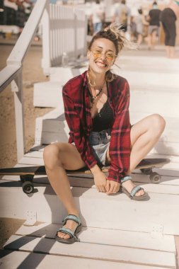 genç gülümseyen kadın güneş gözlüğü ve sahilde ahşap merdivenlerde longboard üzerinde gömlek