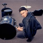 Drummer in black clothing pointing at drum kit on street