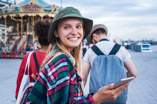 красивая молодая женщина держит смартфон и улыбается перед камерой, проводя время с друзьями в парке развлечений

