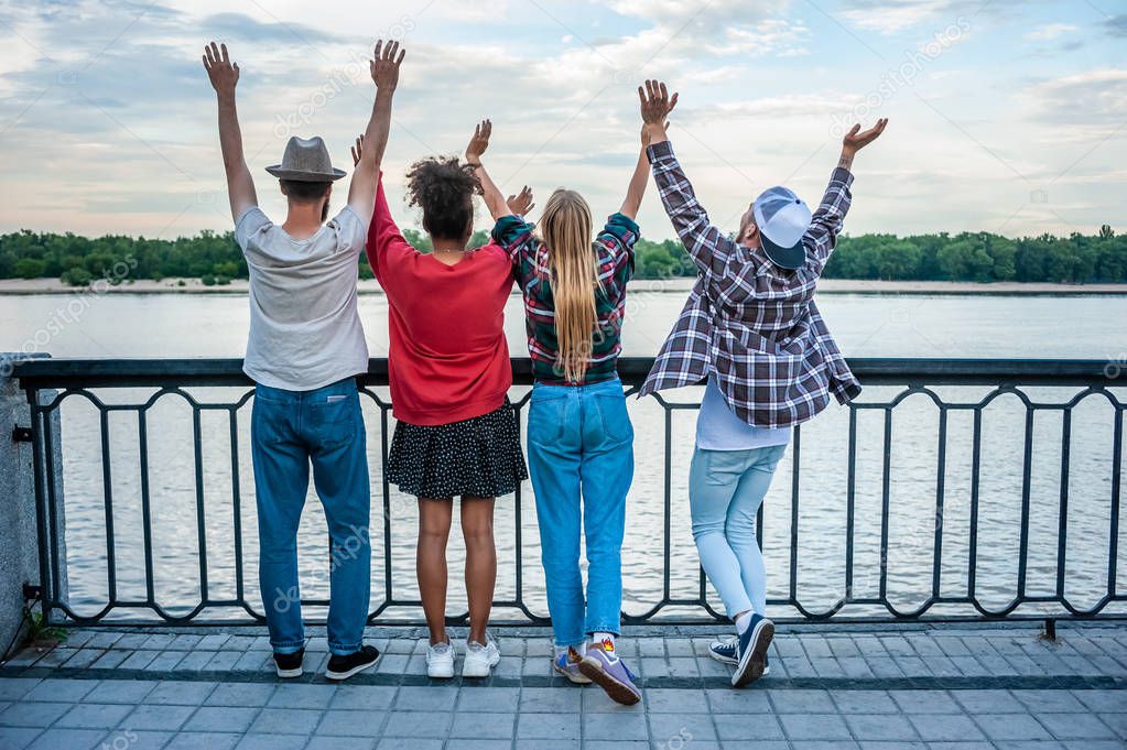 back view of four multiethnic friends raising hands at riverbank