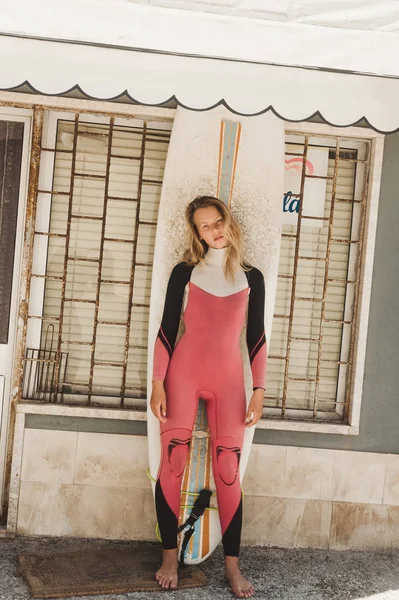 Mujer joven en traje de neopreno con tabla de surf de pie contra la pared de la construcción en Portugal - foto de stock