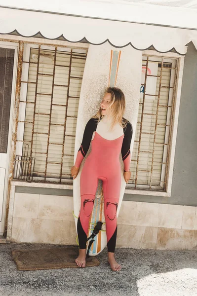Fröhliche Frau im Neoprenanzug mit Surfbrett steht gegen Hauswand in Portugal — Stockfoto