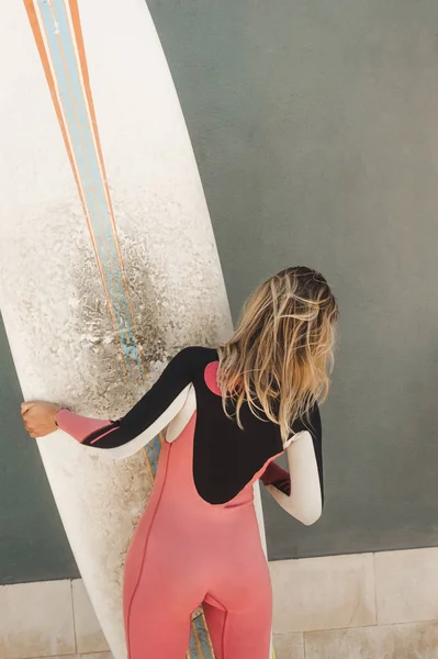 Visão traseira da mulher em roupa de mergulho com prancha de surf contra a parede cinza — Fotografia de Stock