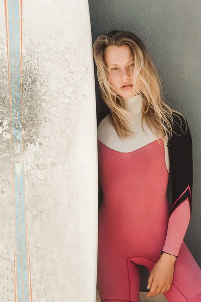 Портрет привлекательной молодой женщины в гидрокостюме с доской для серфинга, стоящей напротив серой стены — стоковое фото