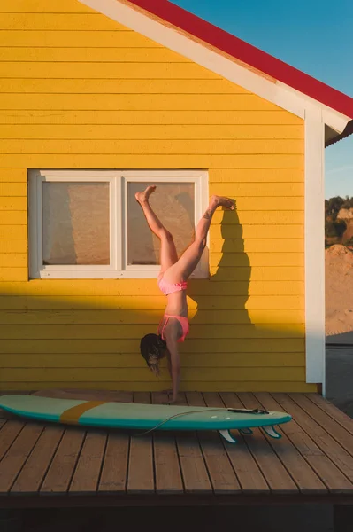 Mujer joven en bikini rosa de pie en las manos contra el edificio amarillo con tabla de surf cerca en Portugal - foto de stock