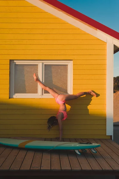 Jeune femme en bikini rose debout sur les mains contre le bâtiment jaune avec planche de surf à proximité au Portugal — Photo de stock