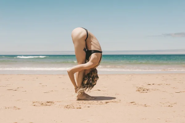 Vista lateral da jovem mulher de pé na postura de ioga de dobra para a frente na praia arenosa — Fotografia de Stock