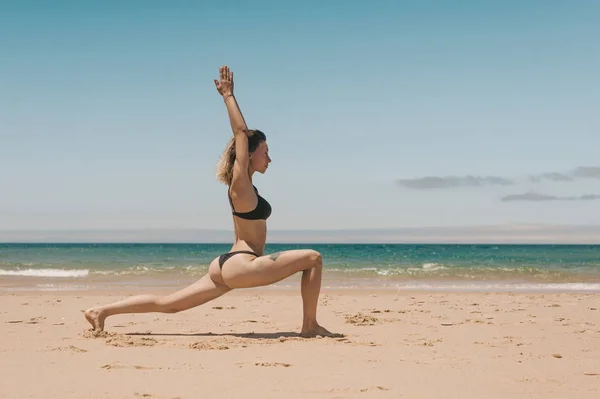 Вид сбоку на молодую женщину в черном бикини, стоящую в позе Воина йоги на песчаном пляже — стоковое фото