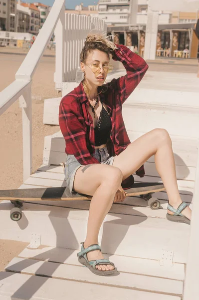 Молодая стильная женщина в солнцезащитных очках и рубашке отдыхает на длинной доске на деревянной лестнице на пляже — стоковое фото