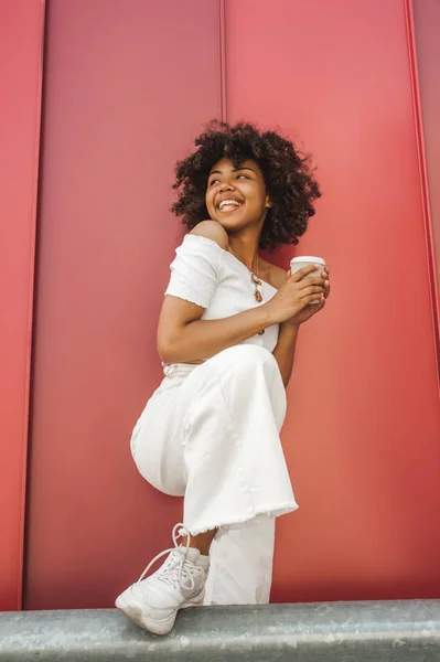 Vista de ángulo bajo de la chica afroamericana feliz sosteniendo café para ir y mirar hacia otro lado - foto de stock