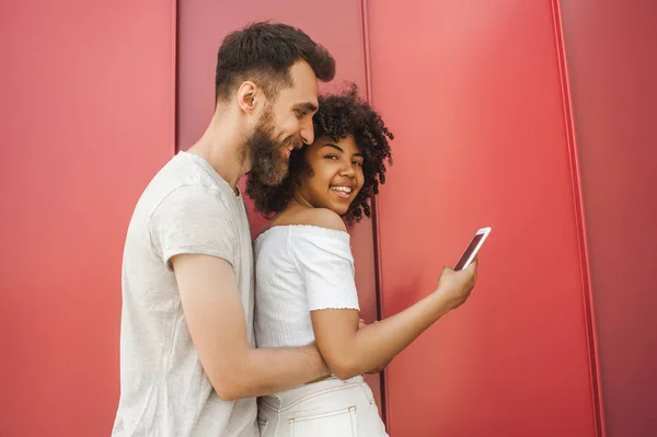 Vista lateral de la feliz pareja joven multicultural utilizando el teléfono inteligente en la calle - foto de stock