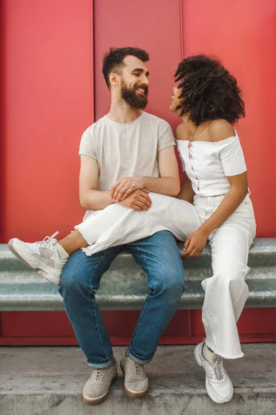 Hermosa feliz joven interracial pareja sonriendo el uno al otro mientras sentado juntos en la calle - foto de stock