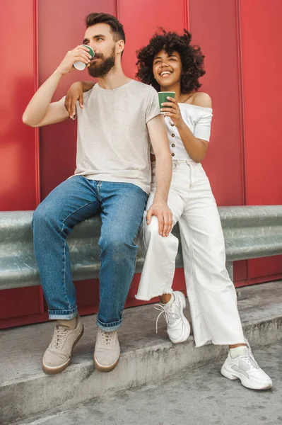 Full length view of stylish young interracial couple drinking coffee from paper cups on street — Stock Photo