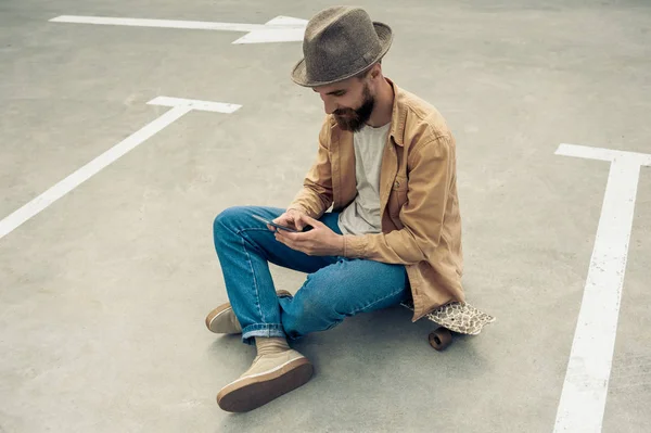 Visão de alto ângulo de homem barbudo elegante sentado no skate e usando smartphone — Fotografia de Stock