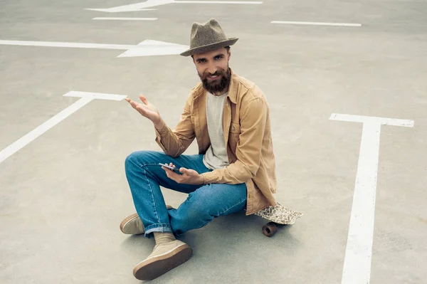 Handsome bearded man looking at camera while sitting on skateboard and using smartphone — Stock Photo