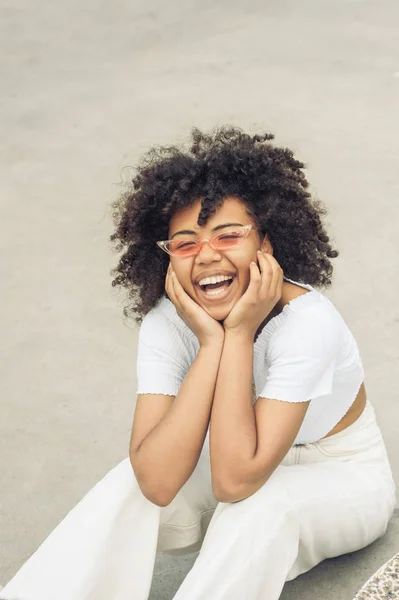 Blick aus der Vogelperspektive auf eine fröhliche junge Afroamerikanerin mit Sonnenbrille, die auf der Straße sitzt und lacht — Stockfoto