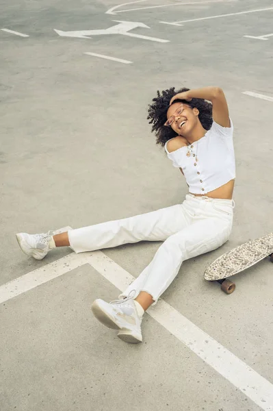 Vista ad alto angolo di allegra ragazza afroamericana seduta vicino allo skateboard e ridendo per strada — Foto stock