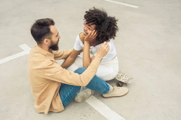 Vista ad alto angolo di felice giovane coppia multietnica sorridente l'un l'altro mentre seduti insieme sulla strada — Foto stock