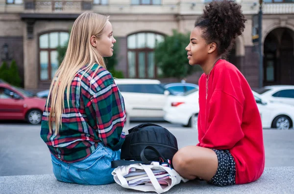 Vista lateral de dos chicas multiétnicas sentadas y mirándose en la calle - foto de stock