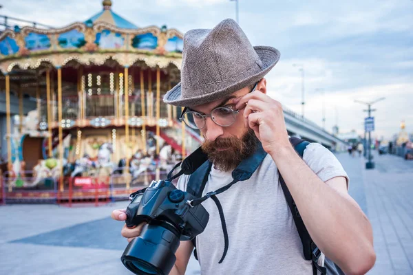 Jovem barbudo de chapéu e óculos usando câmera digital no parque de diversões — Fotografia de Stock