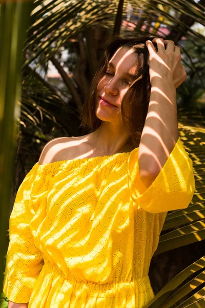美丽的年轻女孩穿着黄色的礼服摆在棕榈叶下 — 图库照片