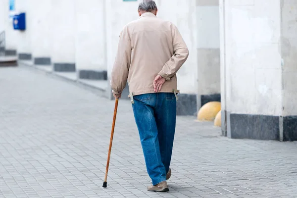 an elderly man with a cane in his left hand walks along the street and holds his right hand behind a sore back