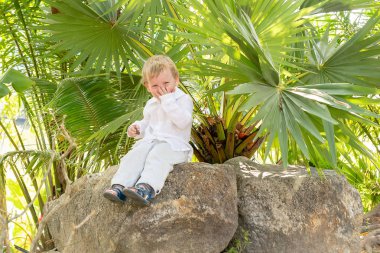 bir taş tropikal ağaçların altında oturan Beyaz giysili küçük sarışın çocuk