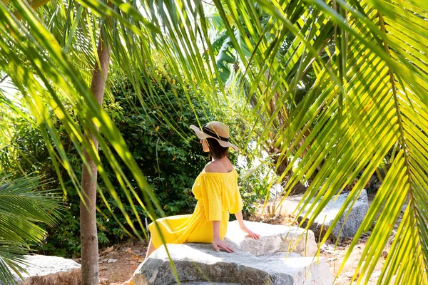 Una joven con un vestido amarillo y sombrero se sienta en una gran roca bajo una palmera — Foto de Stock
