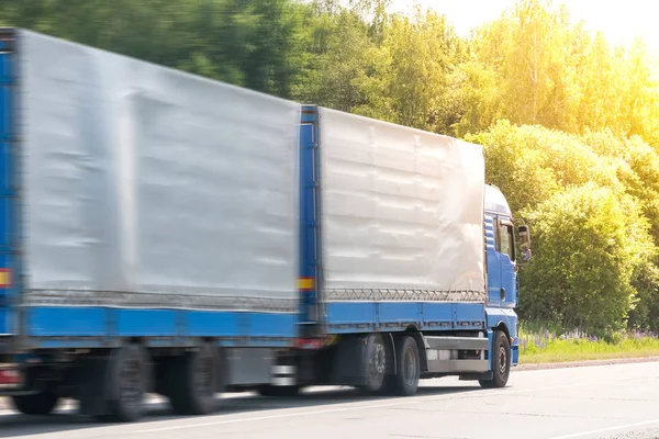 Blue delivery van and white truck driving on the asphalt road between line of trees in autumn landscape — Stock Photo, Image