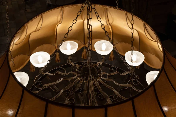 A large antique vintage round metal chandelier of bronze color with decorative elements hangs on the ceiling of a golden background with lighted lamps. — Stock Photo, Image