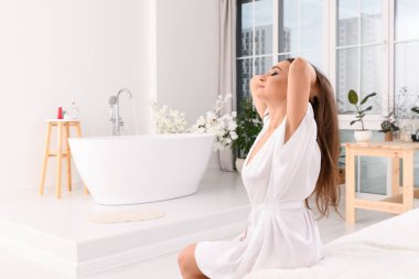 Genç bir kadın beyaz bir bornozla yatağın kenarında oturuyor ve bir kız banyo yapmadan önce saçlarını açıyor. Gözler yana..