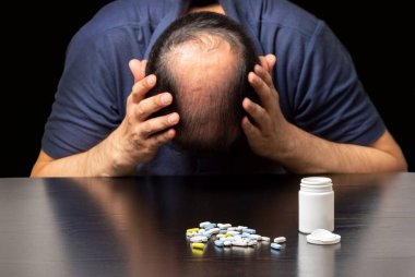 Depresyondaki kel bir adamı, karanlık bir arka planda, masada bir sürü hapın yanına kapat.