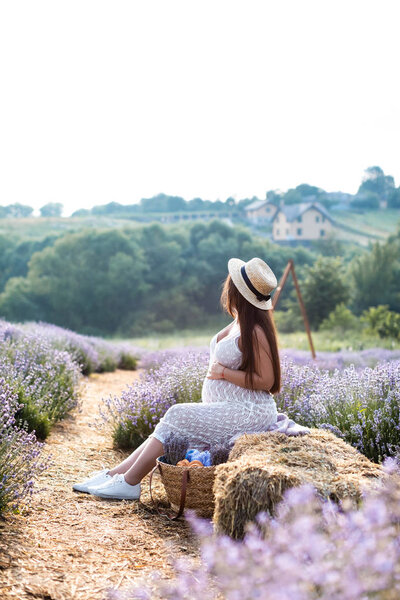 side view of pregnant woman sitting on hay bale in violet lavender field and touching belly