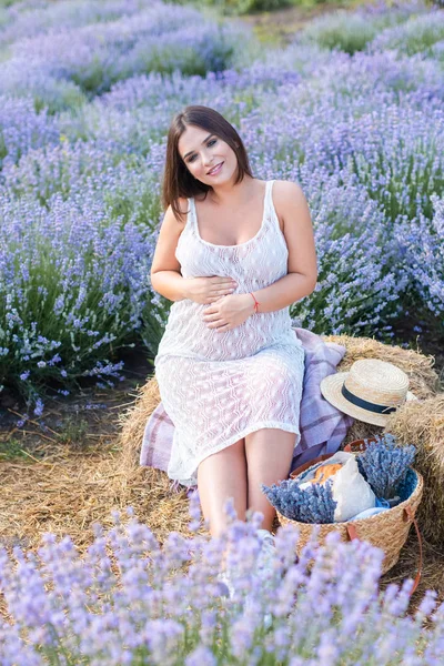 Улыбающаяся беременная женщина сидит на тюке сена на фиолетовом лавандовом поле и трогает живот — стоковое фото
