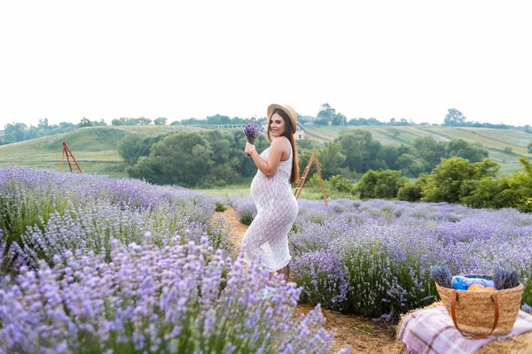 Красива вагітна жінка в білій сукні на фіолетовому лавандовому полі з кошиком для пікніка на тюці — стокове фото