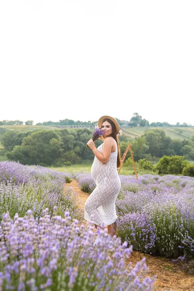 Beautiful smiling pregnant woman in white dress sniffing lavender flowers in field — Stock Photo