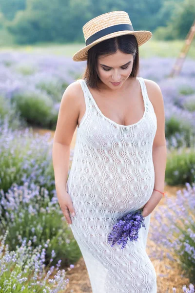 Красивая беременная женщина, стоящая в белом платье на фиолетовом лавандовом поле и держащая маленький букет цветов — стоковое фото