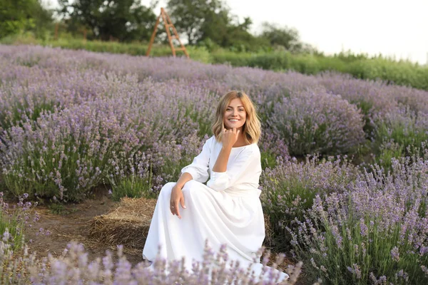 Усміхнена приваблива жінка в білій сукні, що сидить у фіолетовому лавандовому полі — стокове фото