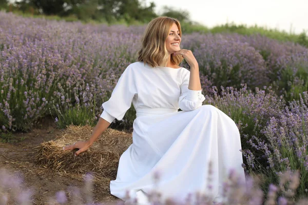 Вид сбоку привлекательной женщины в белом платье, сидящей в фиолетовом лавандовом поле и отводящей взгляд — стоковое фото