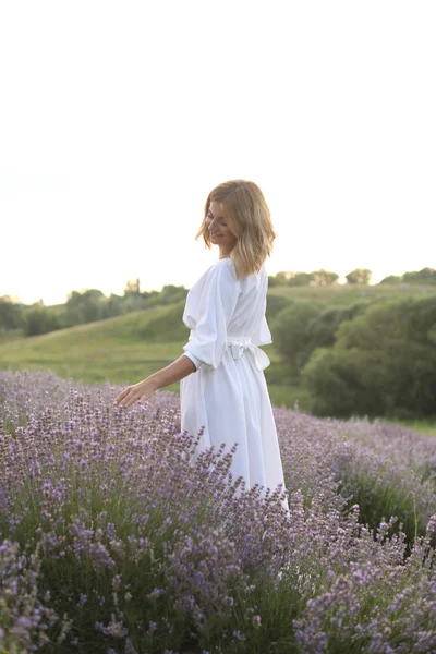 Attraktive Frau in weißem Kleid, die in violettem Lavendelfeld wandelt und Blumen berührt — Stockfoto