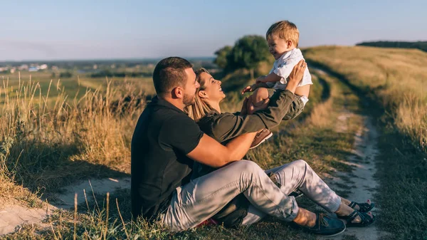 Smiling young parents playing with adorable little son while sitting together on rural trail — Stock Photo