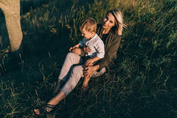 Glückliche junge Mutter blickt in die Kamera, während sie mit ihrem niedlichen kleinen Sohn auf grünem Gras sitzt — Stockfoto