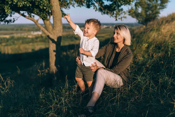 Glückliche junge Mutter sitzt auf dem Gras und schaut ihren niedlichen kleinen Sohn an, der mit dem Finger nach draußen zeigt — Stockfoto