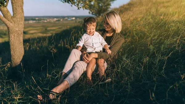 Glückliche junge Mutter und niedlicher kleiner Sohn sitzen zusammen auf grünem Gras bei Sonnenuntergang — Stockfoto