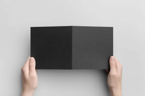 Black Square Bi-Fold Brochure Mock-Up - Male hands holding a black bi-fold on a gray background.