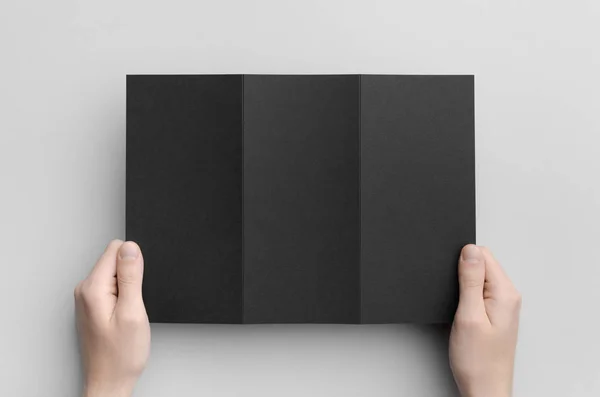 Black A4 Tri-Fold Brochure Mock-Up - Male hands holding a black tri-fold on a gray background.